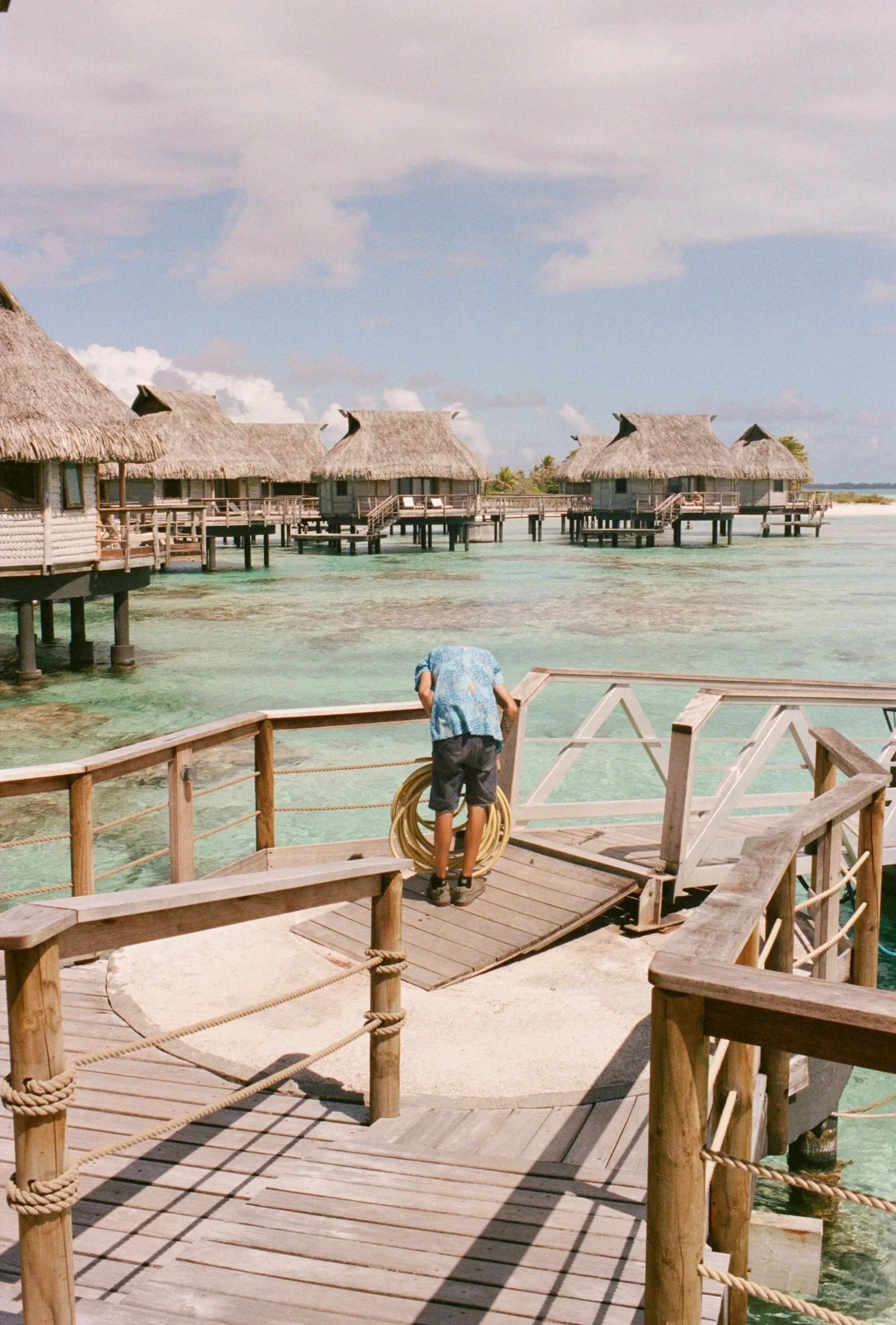 Overwater bungalow resort with wooden walkways above clear turquoise water, overcast sky, and a person in a Hawaiian shirt handling a yellow hose.