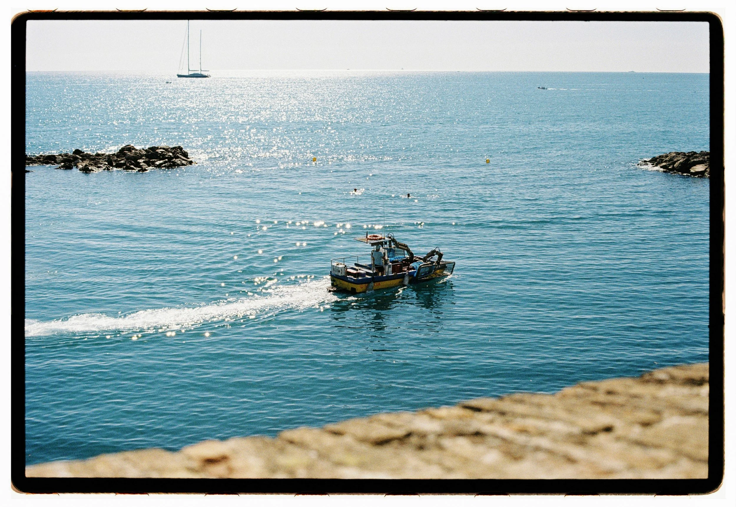 A film photo of a boat sailing in a calm blue ocean near rocky shoreline with a sailboat in the distance under a bright sky.