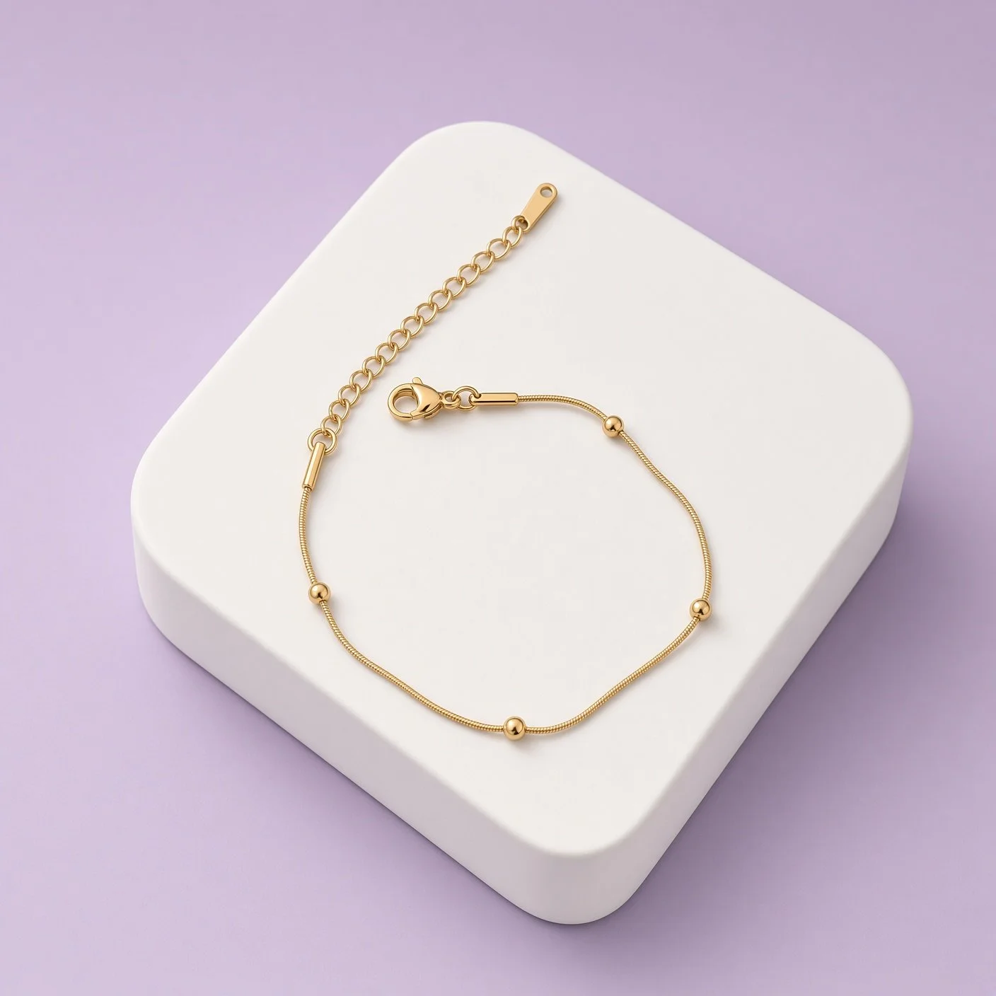 Gold-tone beaded bracelet with chain extender on white display platform, lilac background