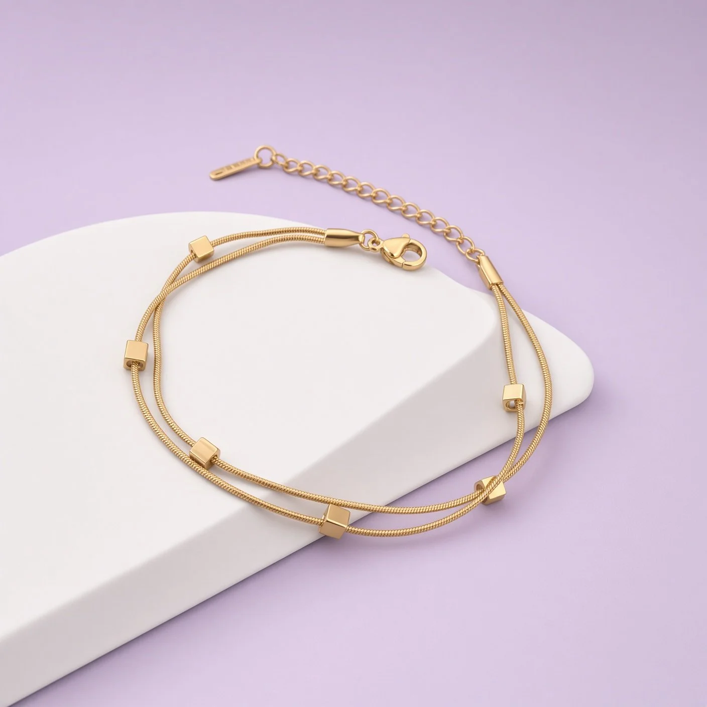 Double-strand gold-tone bracelet with cube charms and extender on white curved stand, lilac