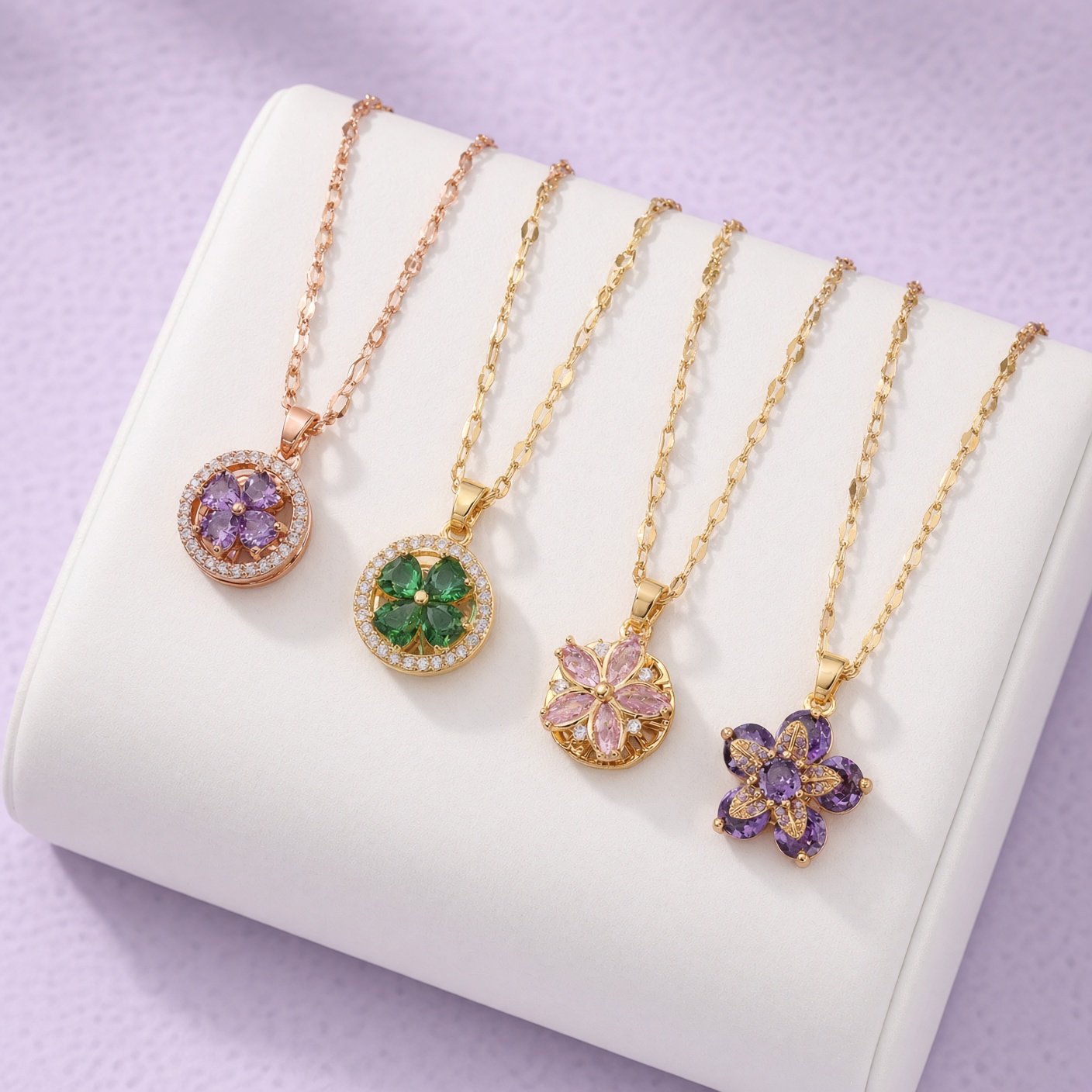 Four gold necklaces with purple, green, and pink flower pendants on white stand