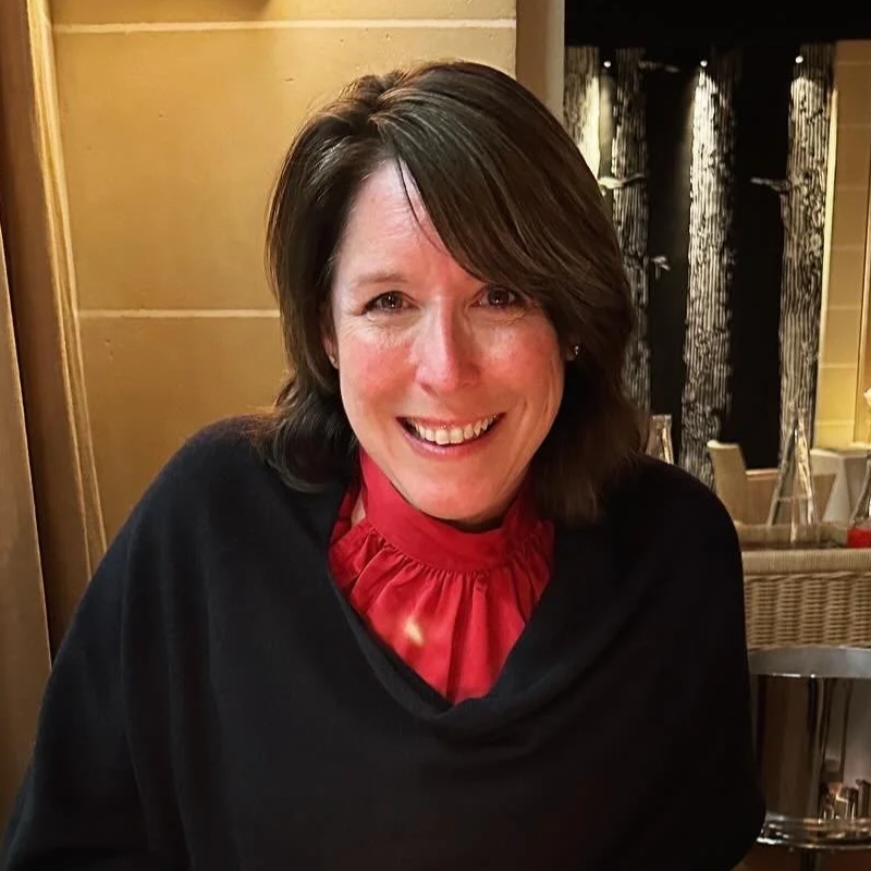 A woman with shoulder-length brown hair smiling at the camera, wearing a black top and a red ruffled collar, sitting in a restaurant with a warm-toned interior.