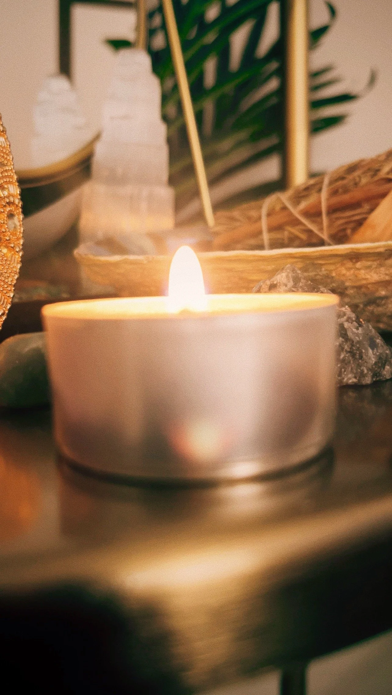 A lit tealight candle in a round metal holder on a table with decorative objects, plants, and natural elements in the background.