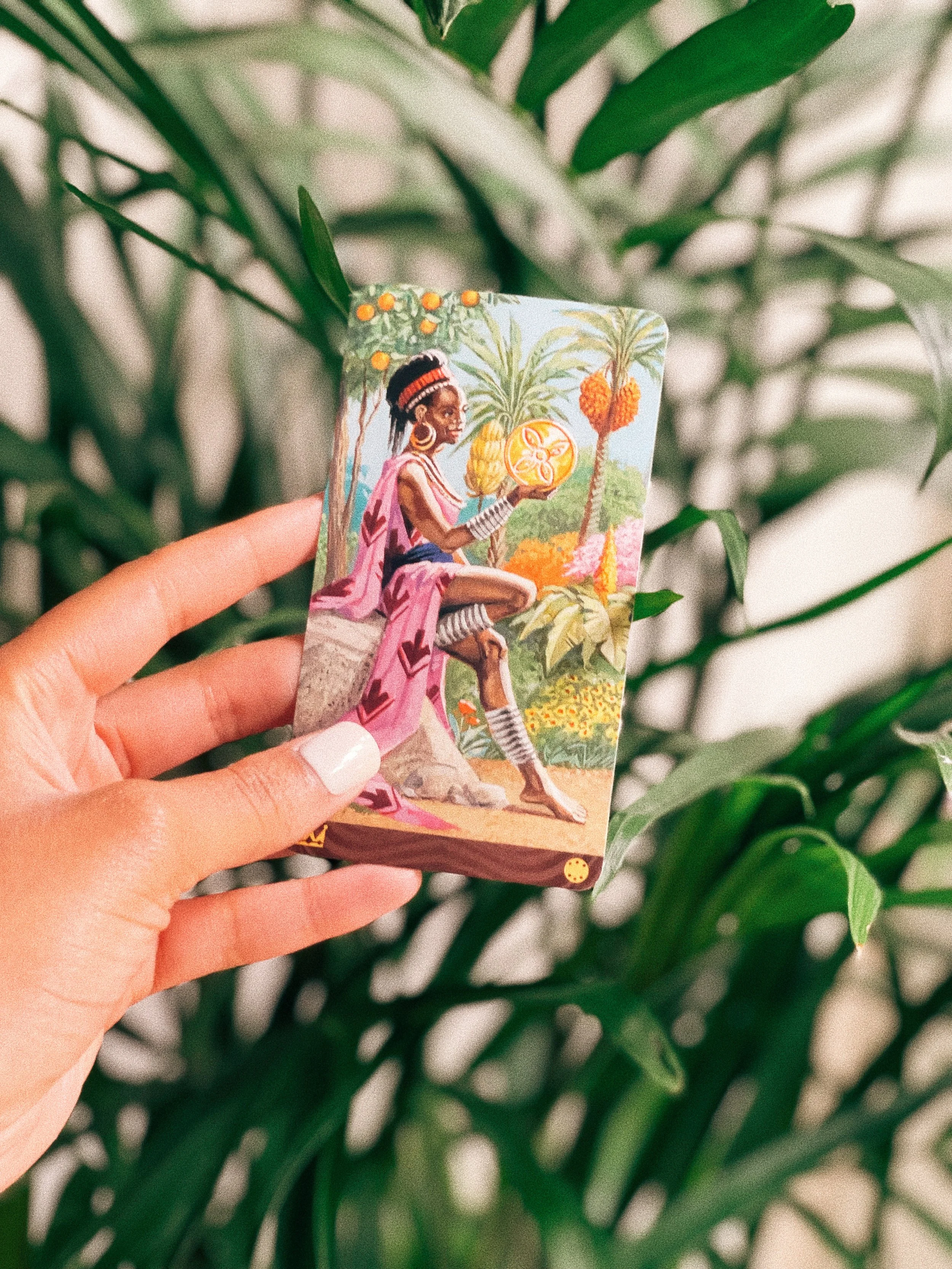 A Queen of Pentacles tarot card featuring an African woman in traditional clothing holding a large coin, with lush tropical plants in the background.
