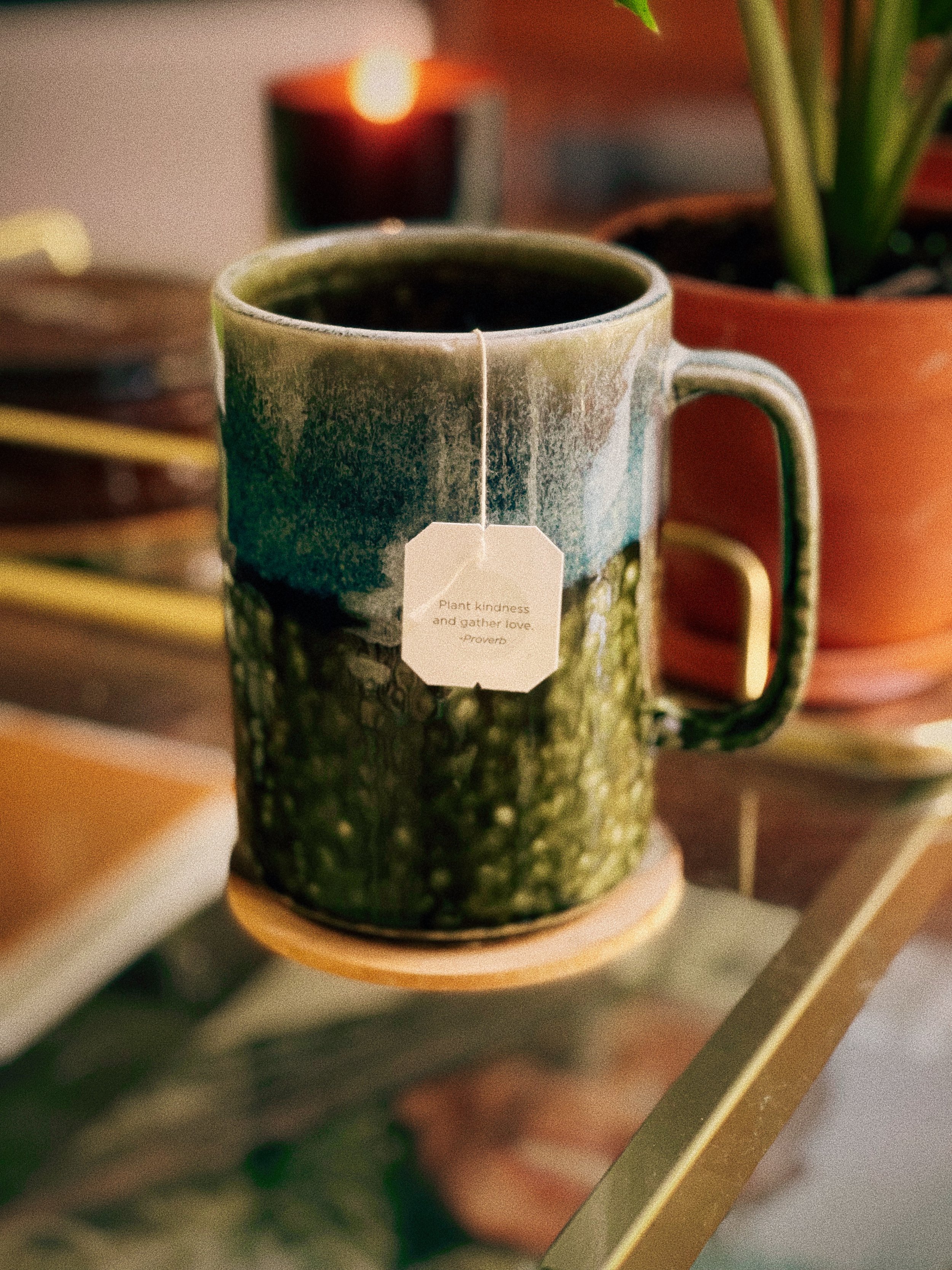 A ceramic mug filled with tea, resting on a wooden coaster on a glass table, with a potted plant and a candle in the background.