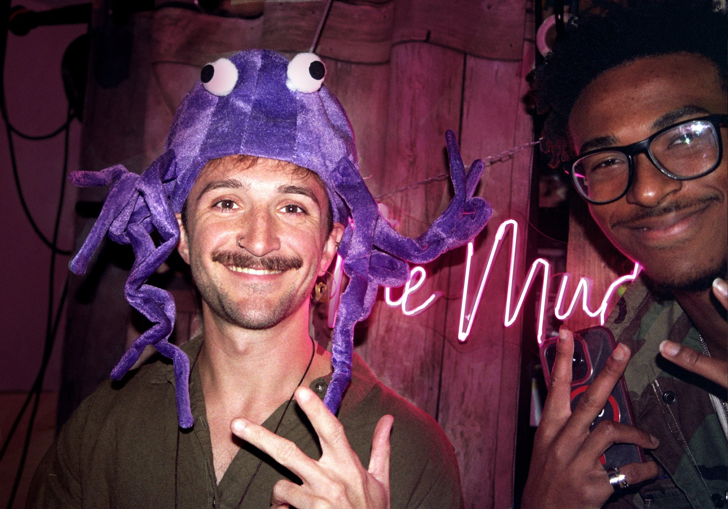 Two men smiling at the camera, one wearing a purple octopus hat, and the other wearing glasses. They are making peace signs with their hands in front of a pink neon sign that says "le Mur." The background features a wooden wall.