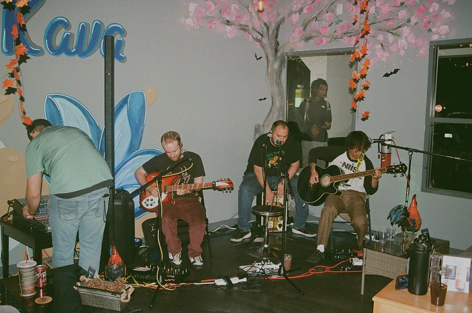 A band performing live indoors with four members playing guitars and singing, surrounded by colorful wall art including a pink cherry blossom tree, a blue butterfly, and bats.