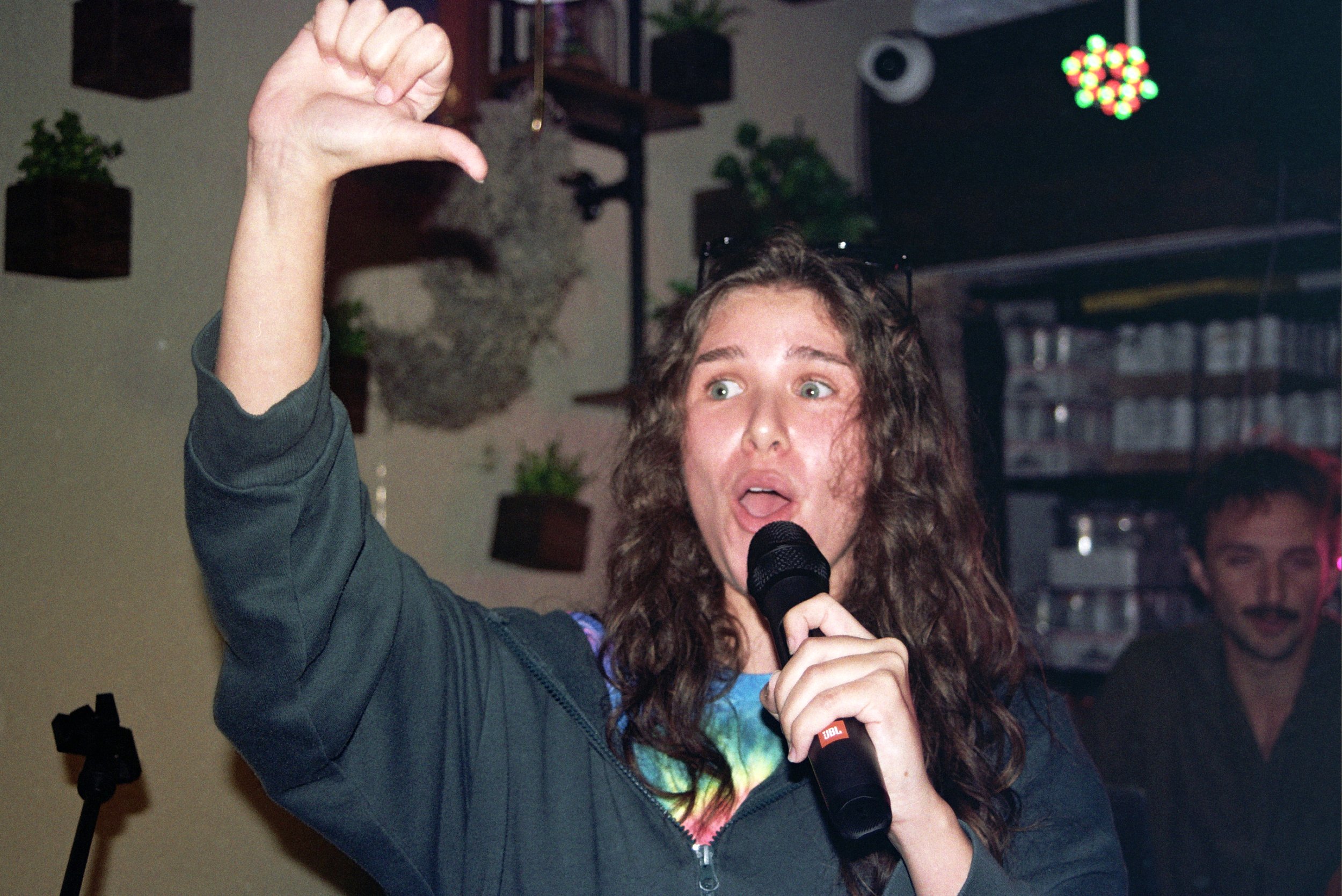 A young woman with long wavy hair holding a microphone in her right hand, with her left arm raised and thumb pointing downward, speaking or performing in an indoor setting with shelves and plants in the background.