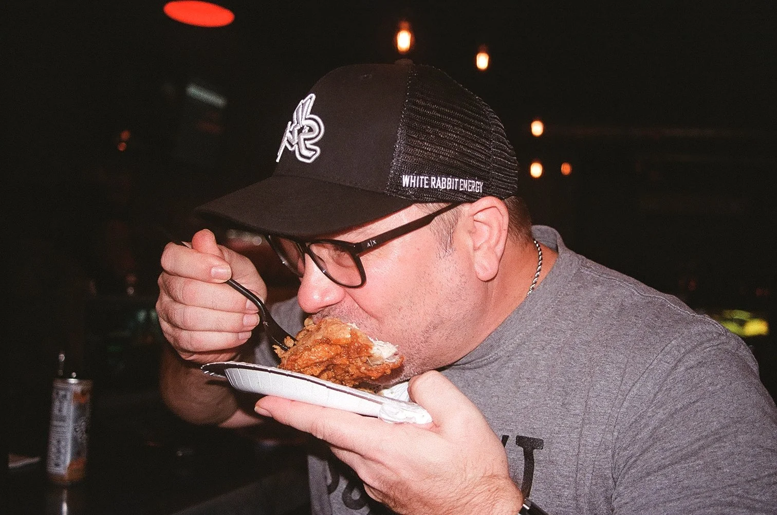 A man wearing glasses and a black cap with white text, eating fried chicken from a paper plate in a dimly lit setting.