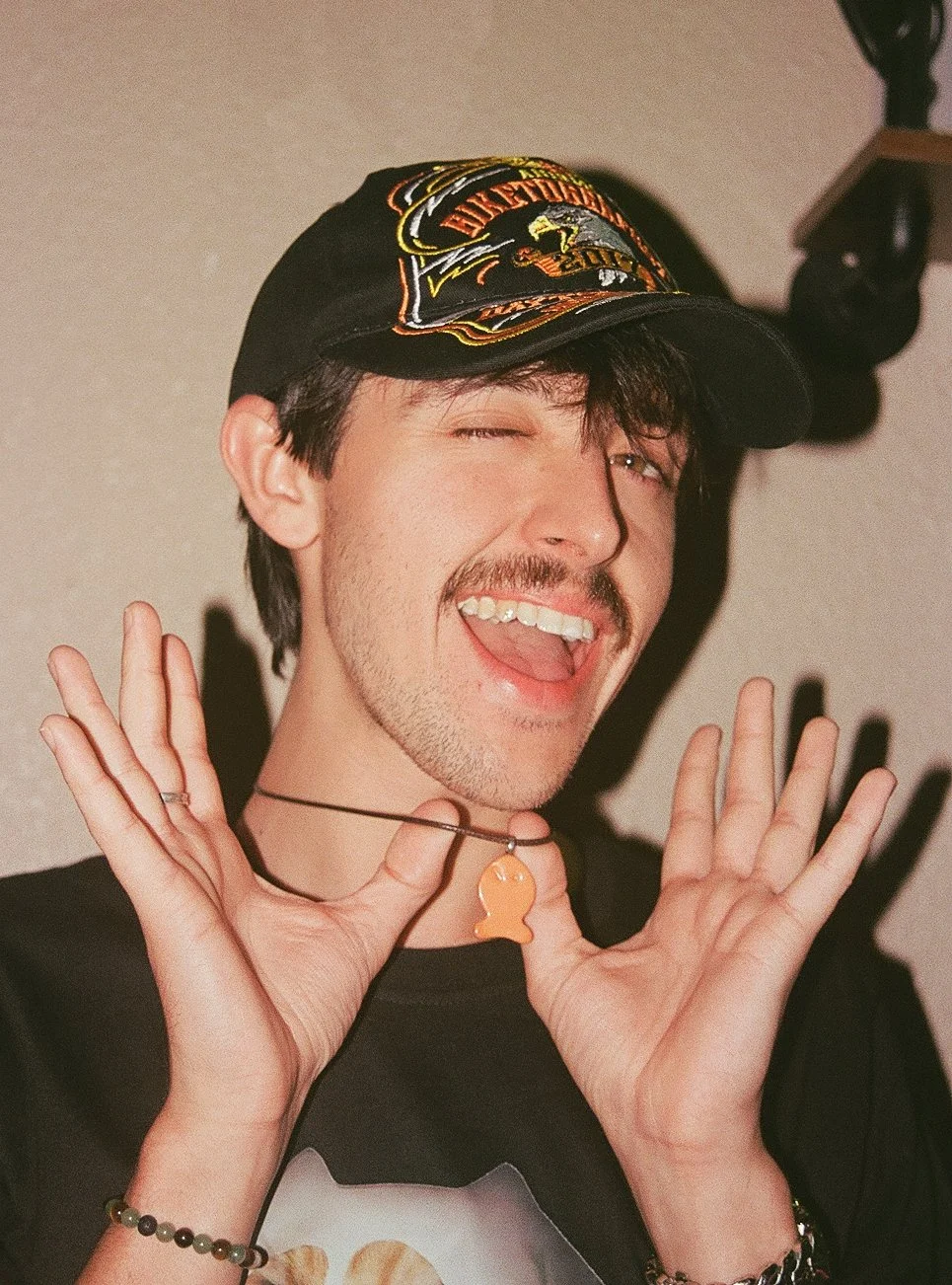 A young man with dark hair, a mustache, and a beard is wearing a black cap, a necklace with an orange fish-shaped pendant, and a black T-shirt. He is smiling with his mouth open and winking, holding his hands near his face with fingers spread.