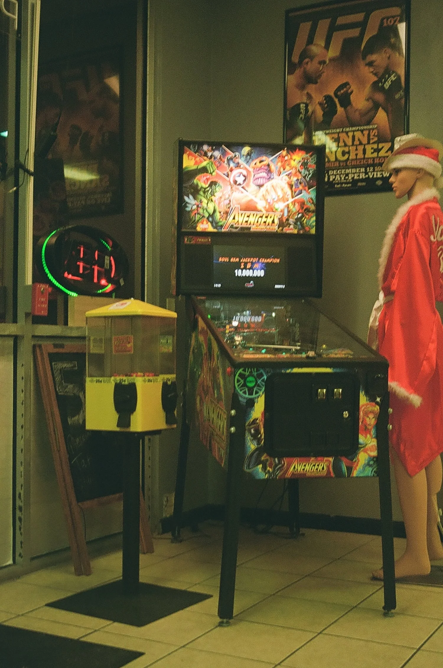 An indoor arcade featuring a Marvel Avengers-themed pinball machine, a UFC poster on the wall, a mannequin dressed as Santa Claus, and a yellow donation or toy machine. The setting appears to be during the holiday season.