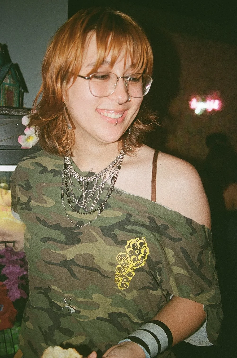 A young woman with shoulder-length reddish hair, glasses, and many layered necklaces smiling while looking down. She wears a camouflage shirt with a yellow graphic of a revolver on it over a strap top. She has a nose ring and labret piercing, and a s