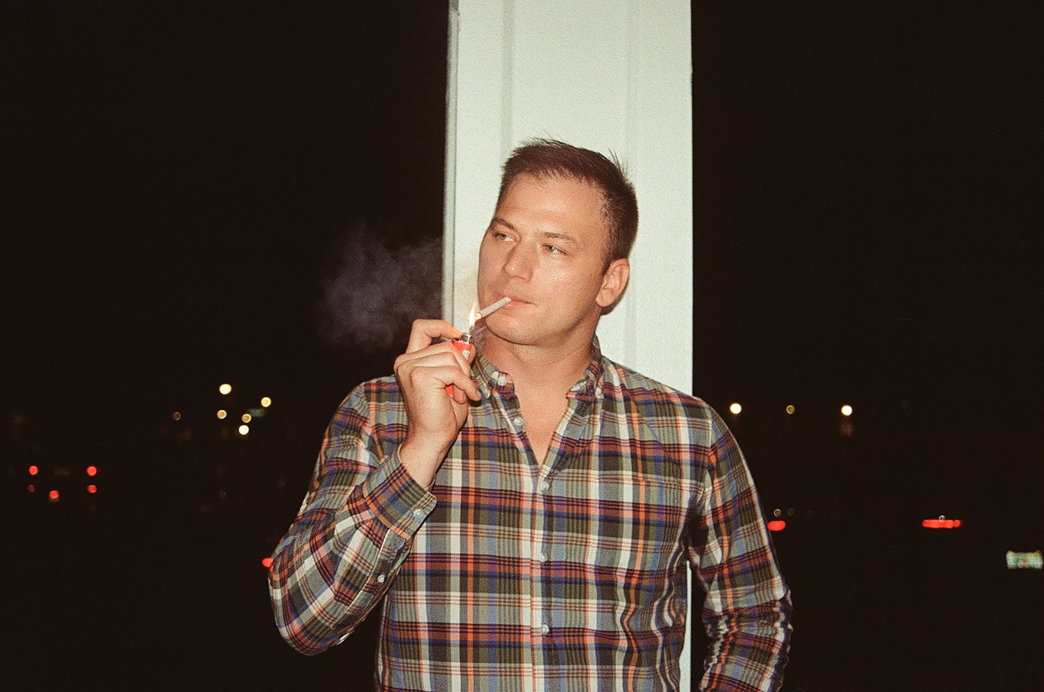 A man in a plaid shirt standing outside at night, smoking a cigarette with smoke rising, against a dark background with distant city lights.