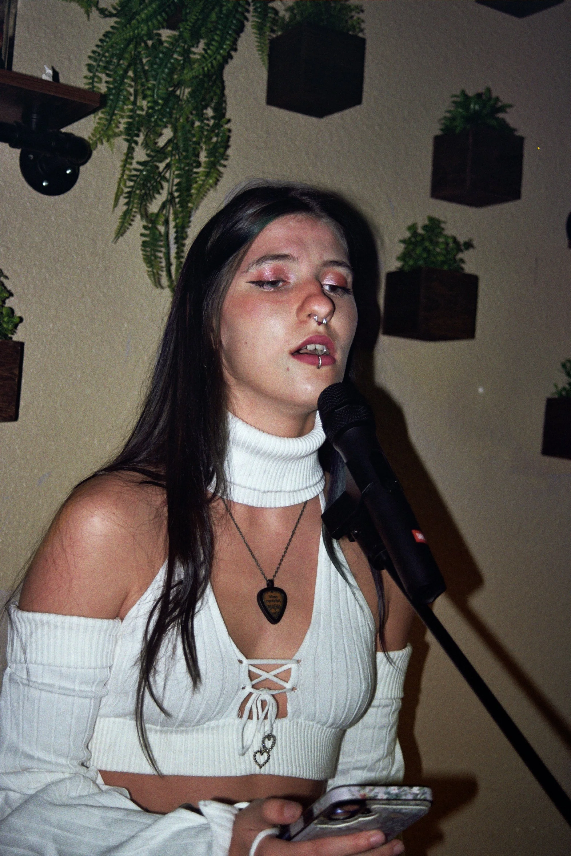 A young woman with dark hair, wearing a white off-shoulder top with a lace-up front and a white turtleneck underneath, is singing or speaking into a microphone. She has piercings and wears necklaces, one with a guitar pick pendant. She is holding a s