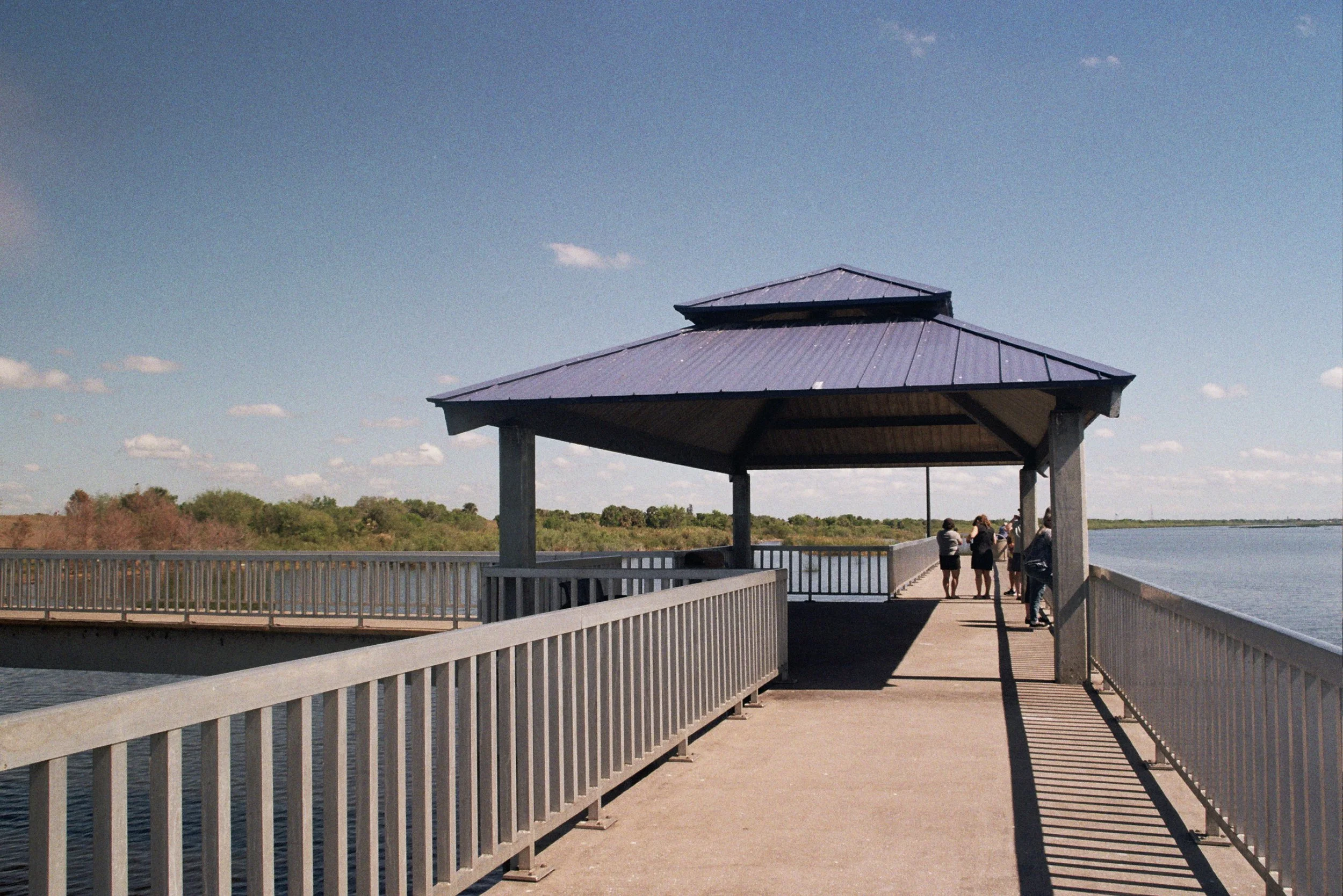 A scenic view of a pier with a pavilion over the water, with people walking and enjoying the view, and trees visible in the background under a partly cloudy sky.