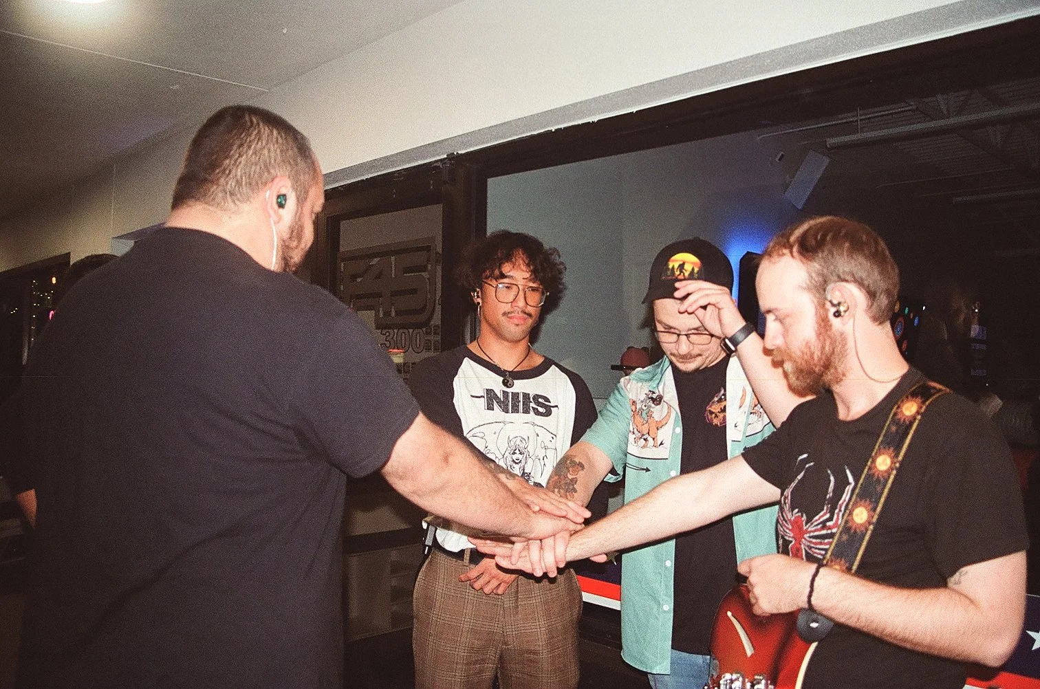 Four men with light skin standing close together holding hands in a circle in a dimly lit room, with one playing a guitar. The man on the far left is seen from behind, the second man has curly hair and glasses, the third man is wearing a cap and glas