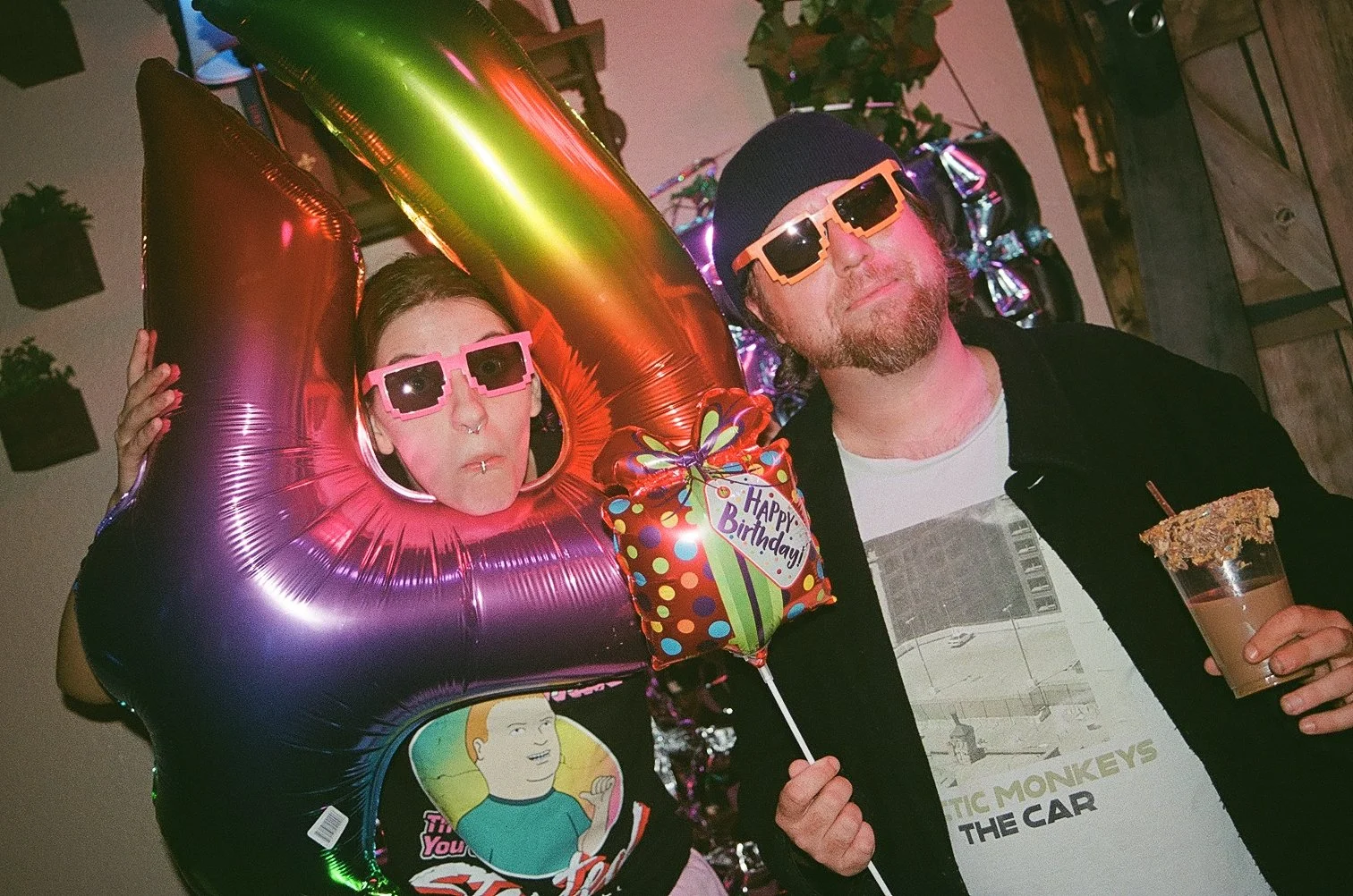 Two people celebrating a birthday. The woman is wearing pink sunglasses and a birthday hat, blowing a party blower, and holding a 'Happy Birthday' balloon. The man is wearing yellow sunglasses, a black beanie, and a graphic t-shirt, holding a drink with a sugared rim.