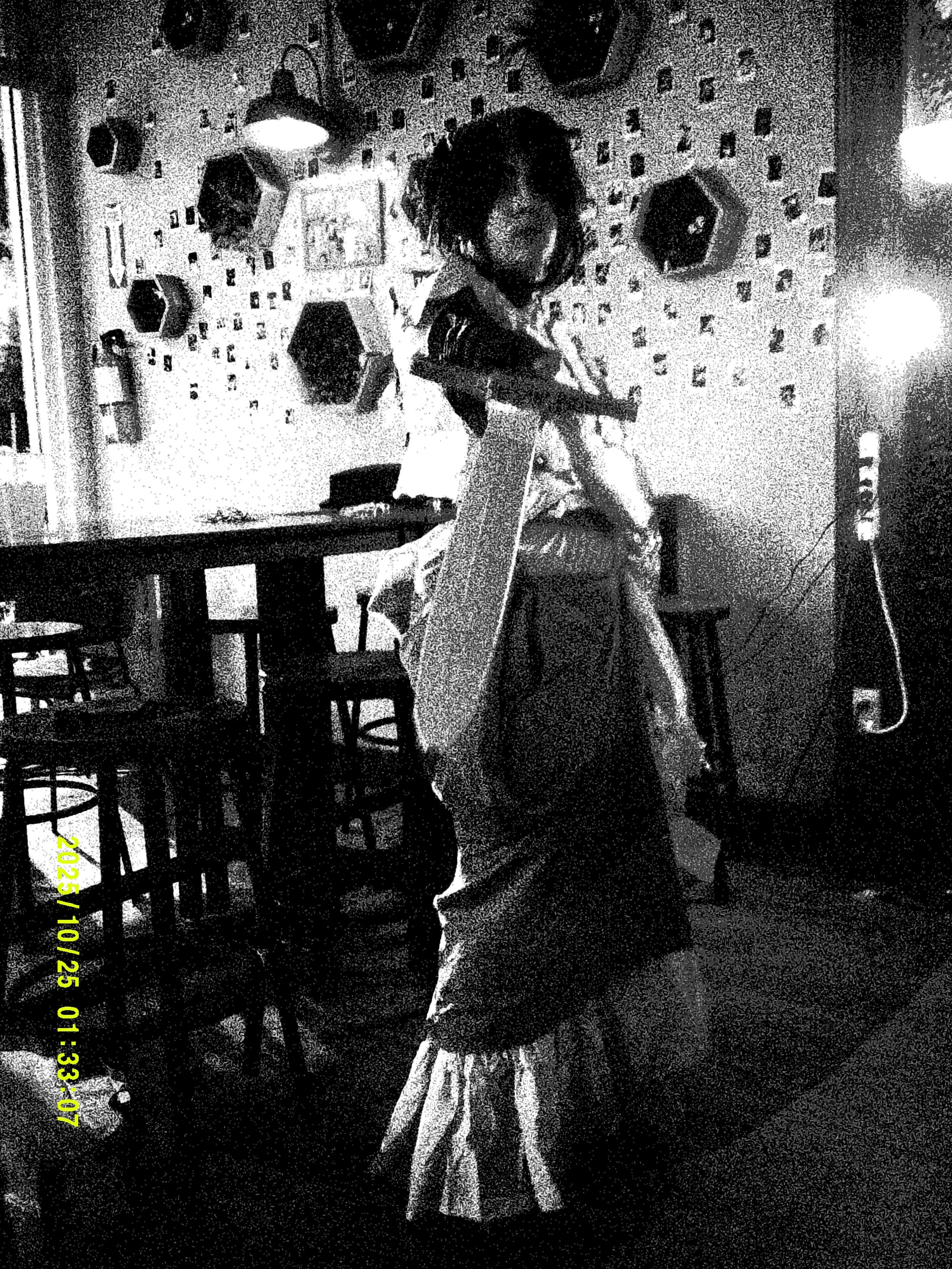 A woman with curly hair is mid-dance in a dimly lit room, wearing an apron and a long skirt. The background has wall decorations, shelves, a fire extinguisher, and tables and chairs.