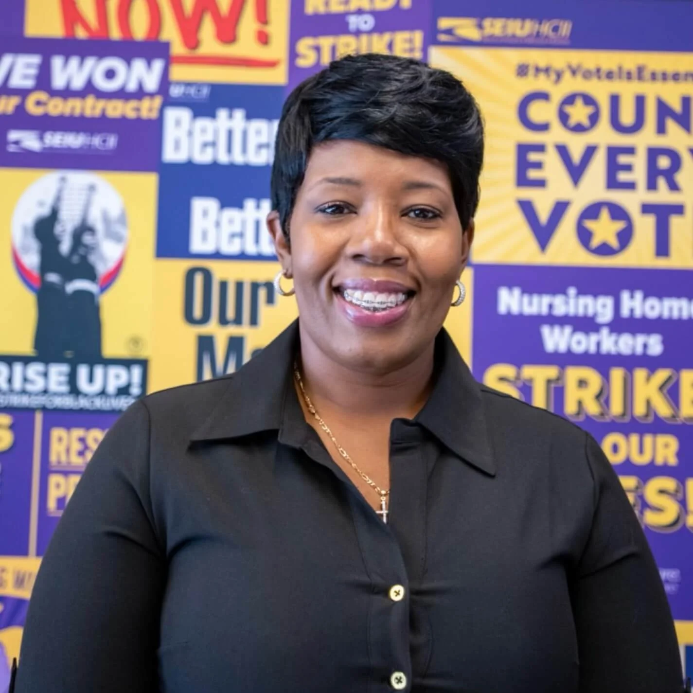 A middle-aged African American woman with short black hair, earrings, and a gold necklace, smiling in front of a colorful background with various election-related posters and slogans in yellow, purple, and white.