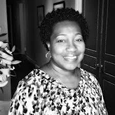 A smiling African American woman with short, curly hair, wearing a patterned top, standing in a room with dark furniture.