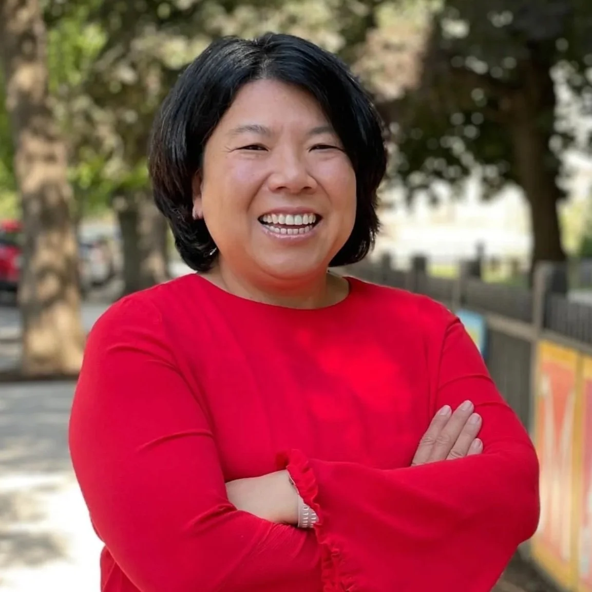 Middle aged Asian woman with short black hair wearing a red top, standing outdoors while smiling with trees and a fence in the background.
