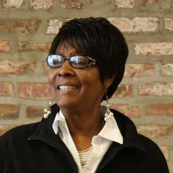 A smiling African American woman with short black hair and glasses, wearing a white collared shirt and black jacket, standing in front of a brick wall.