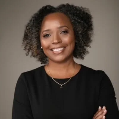 Young African American woman with natural curly hair, smiling, wearing a black top and a gold necklace, against a neutral background.