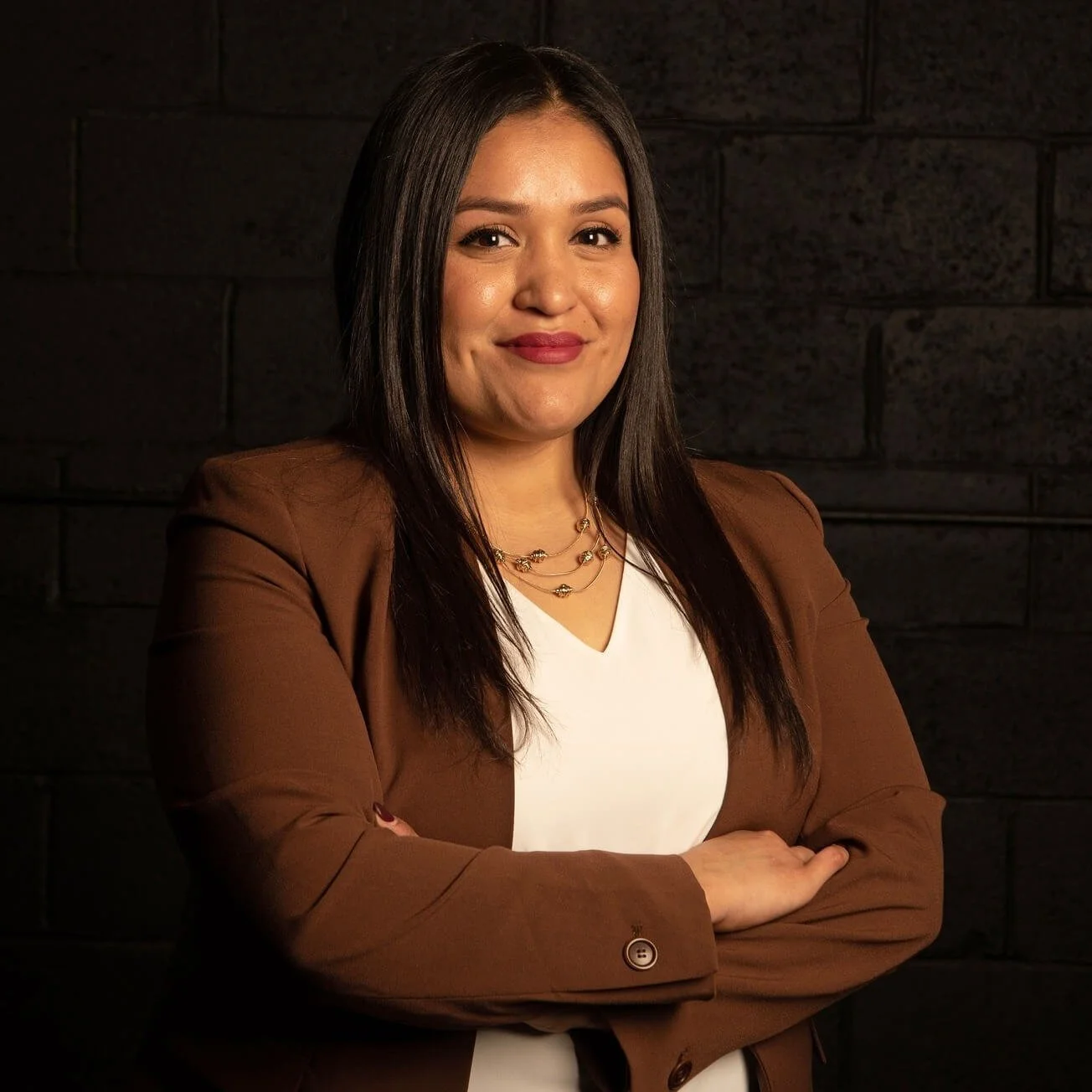 A young Hispanic woman with long dark hair in a brown blazer over a white top, standing with arms crossed, smiling, against a dark brick wall background.