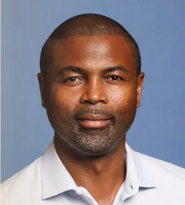 Close-up portrait of a middle-aged African American man with short black hair, neatly trimmed beard, and wearing a white collared shirt against a plain blue background.