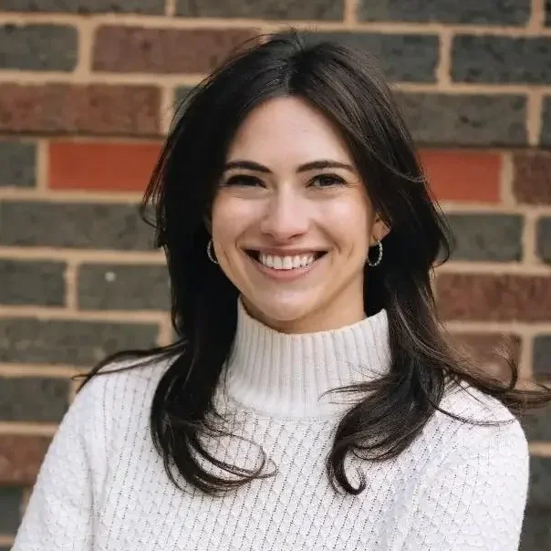 A woman with dark hair smiling in front of a brick wall, wearing a white turtleneck sweater and hoop earrings.
