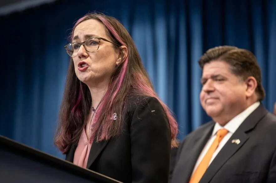 A woman with glasses and long hair with pink streaks speaking at a podium, with a man in a suit and tie standing beside her against a blue curtain background.