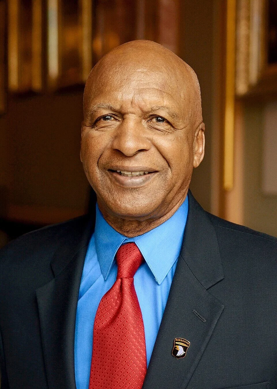 A smiling elderly man in a dark suit, light blue shirt, and red tie, wearing a U.S. Army airborne badge on his lapel, standing indoors.