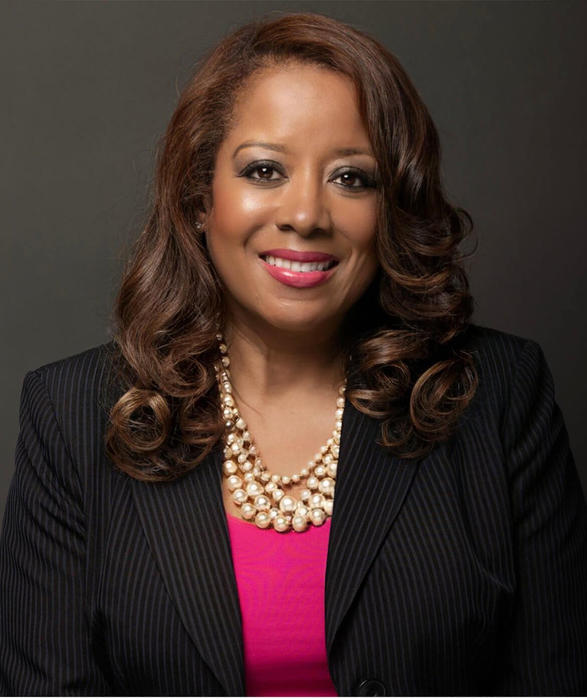 A smiling African American woman wearing a black pinstripe blazer, pink blouse, and pearl necklace, smiling with long curly hair, against a dark background.