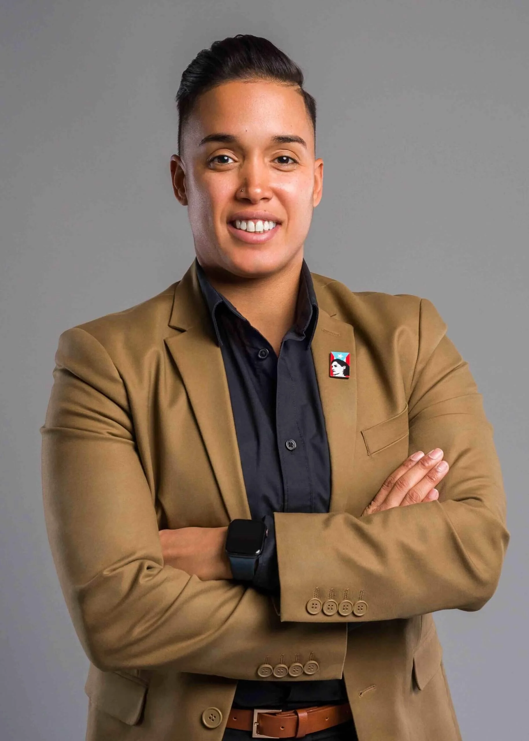 A non-binary person of Hispanic descent with short dark hair, wearing a tan blazer with a small pin, black shirt, and smartwatch, smiling with arms crossed against a gray background.