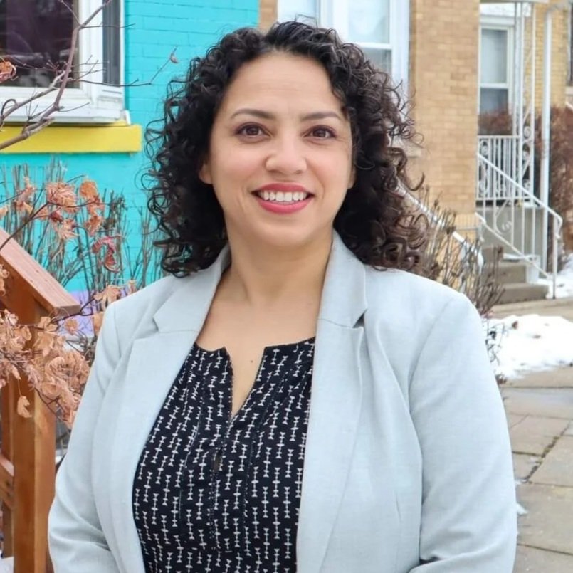 A middle-aged Hispanic woman with curly dark hair smiling, wearing a light gray blazer over a black patterned top, standing outdoors near a blue house with yellow window trim and bushes with dried brown leaves.