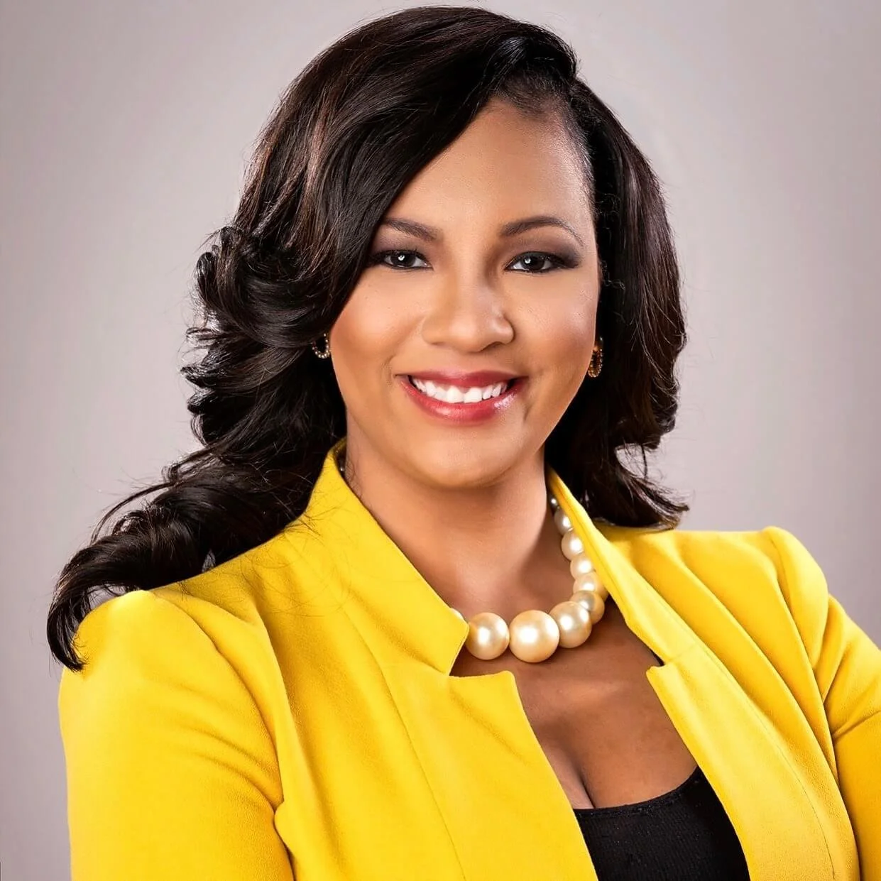 An African American woman with dark wavy hair, wearing makeup, a bright yellow blazer, a string of large pearls, and earrings, smiling against a plain background.