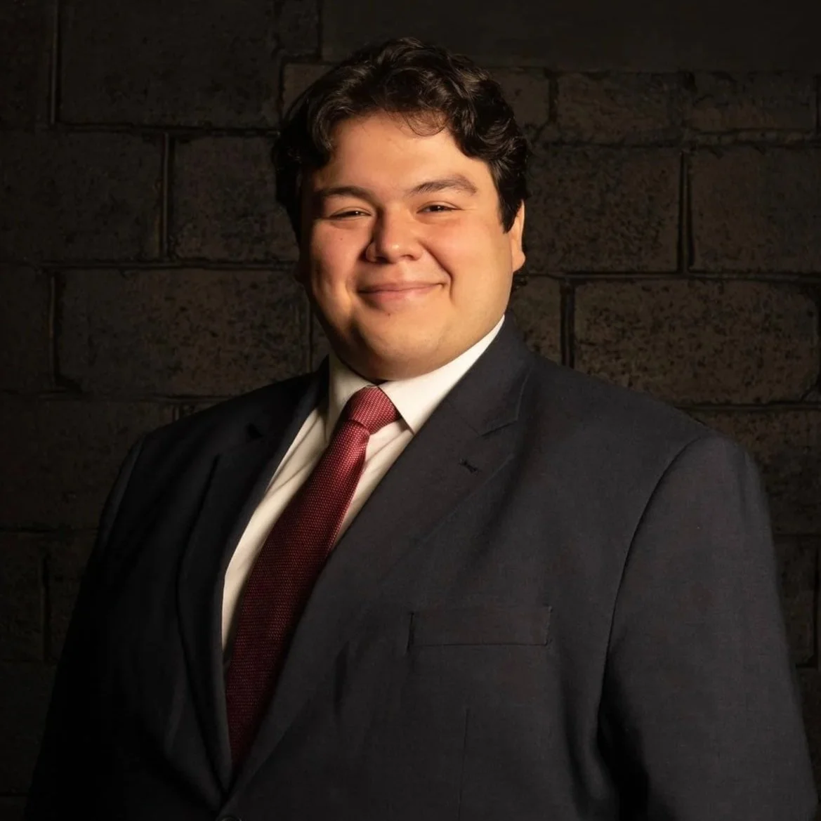 A young Hispanic man in a dark suit with a white shirt and red tie, smiling in front of a dark brick wall.