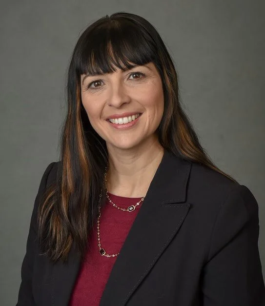 A smiling Hispanic woman with long dark hair and bangs wearing a black blazer over a maroon top, with a silver necklace, against a plain gray background.