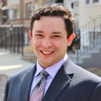 A Hispanic man in a suit and tie smiling outdoors in front of residential buildings.