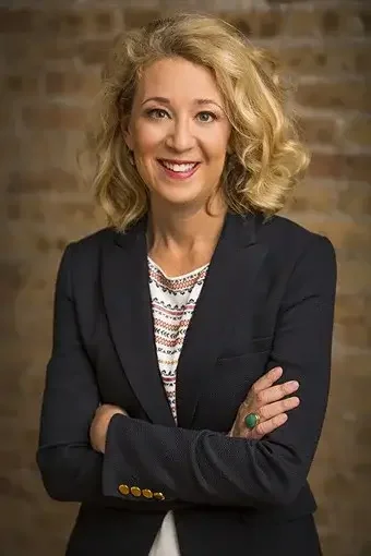 A woman with blonde curly hair smiling, wearing a dark blazer over a patterned blouse, standing against a brick wall background.