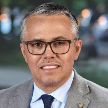 Portrait of a man with gray hair and glasses wearing a gray suit and blue tie, standing outdoors with blurred lights in the background.