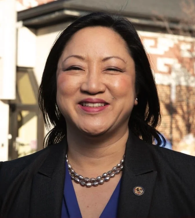 A woman with black hair smiling outdoors, wearing a black blazer, blue top, pearl necklace, and small earrings.
