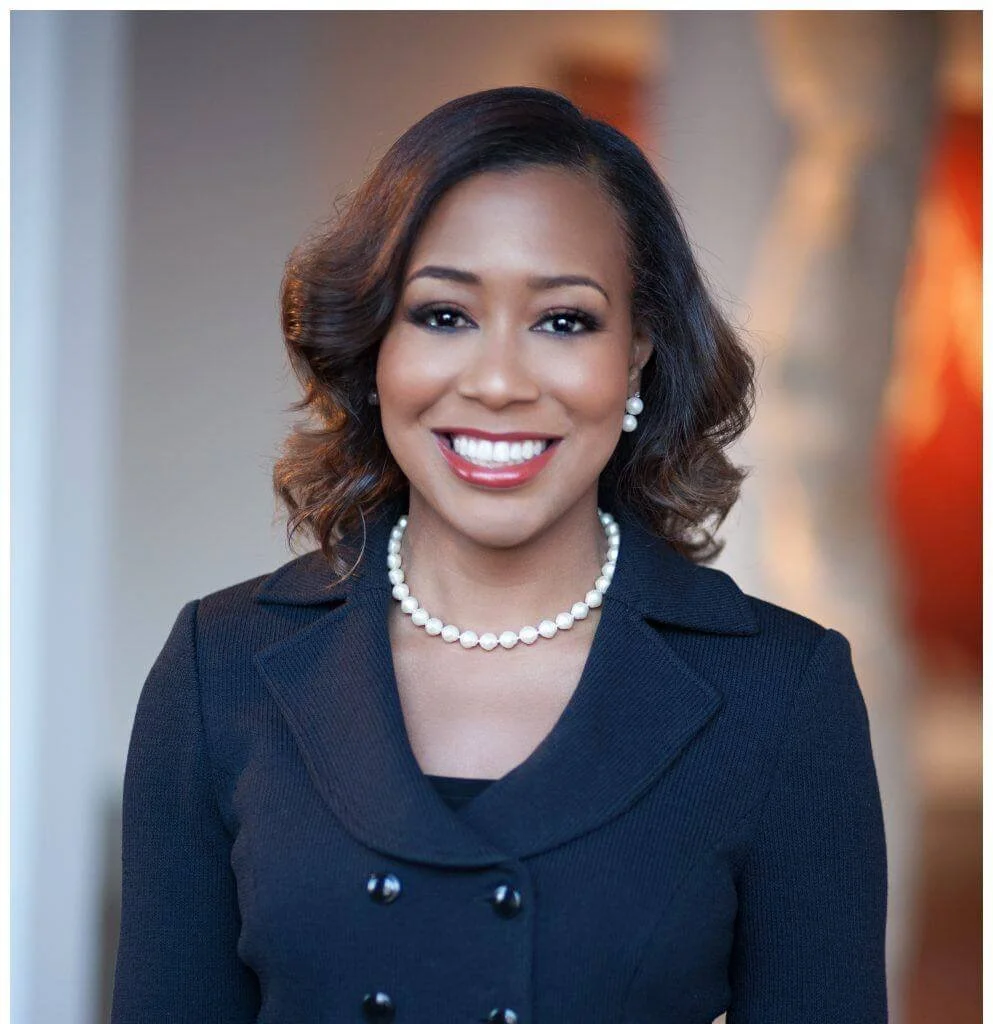 An African American woman with shoulder-length dark hair styled in loose waves, smiling, wearing a navy blazer with a dress underneath, a pearl necklace, and matching pearl earrings, with an blurred indoor background.