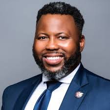 Portrait of a smiling African American man with dark hair and a beard, wearing a dark suit, white shirt, and a tie, against a plain gray background.