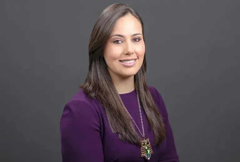 Portrait of a young Hispanic woman with long dark hair, wearing a purple top and a colorful necklace, smiling against a gray background.