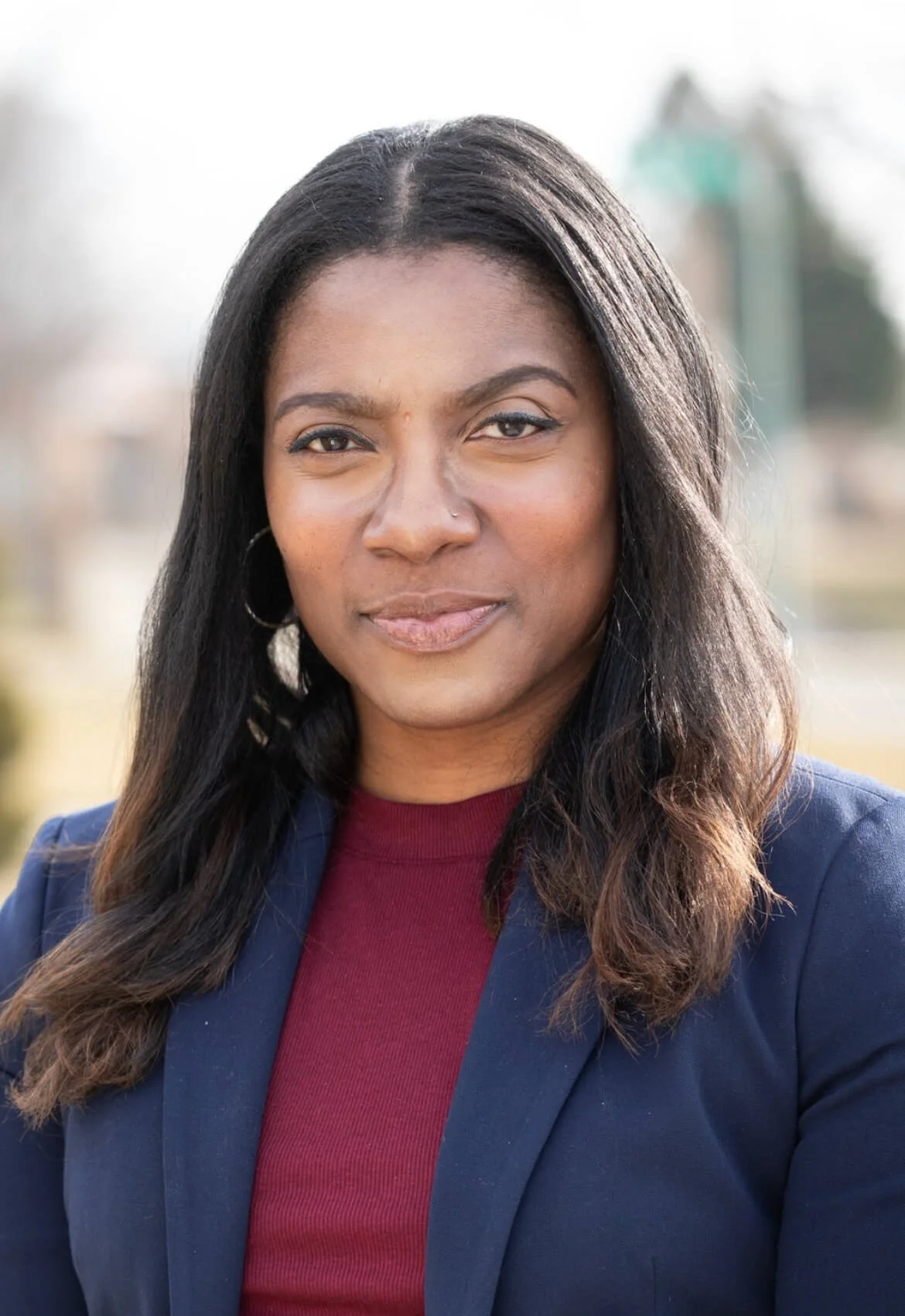 An African American woman with long black hair, wearing a navy blazer and red top, standing outdoors with blurred background.