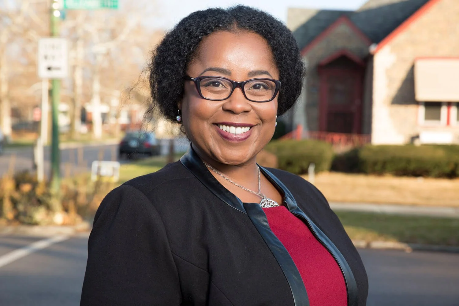 An African American woman with glasses and short curly hair smiling outdoors, wearing a black jacket over a burgundy top, with houses and trees in the background.