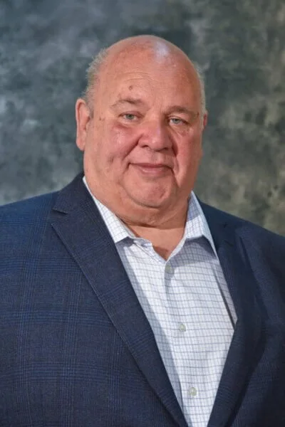 Portrait of a bald man in a blue blazer and white checkered shirt against a mottled gray background.
