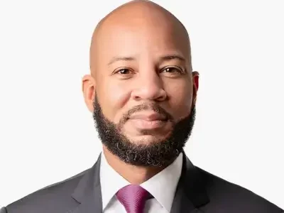 An African American man with a bald head and a beard, wearing a dark suit, white shirt, and maroon tie, looking at the camera with a neutral expression.