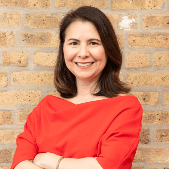 A smiling woman with shoulder-length brown hair, wearing a red dress with an off-the-shoulder neckline, standing against a brick wall.